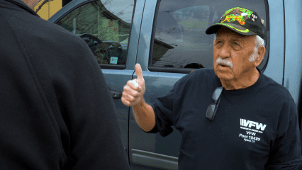 An older man wearing a black VFW shirt and decorated hat smiling and giving a thumbs up next to a parked van, expressing approval or satisfaction.