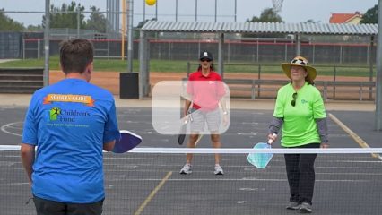 A youth and two women are playing pickleball outdoors. Image also includes a video play button overlay.