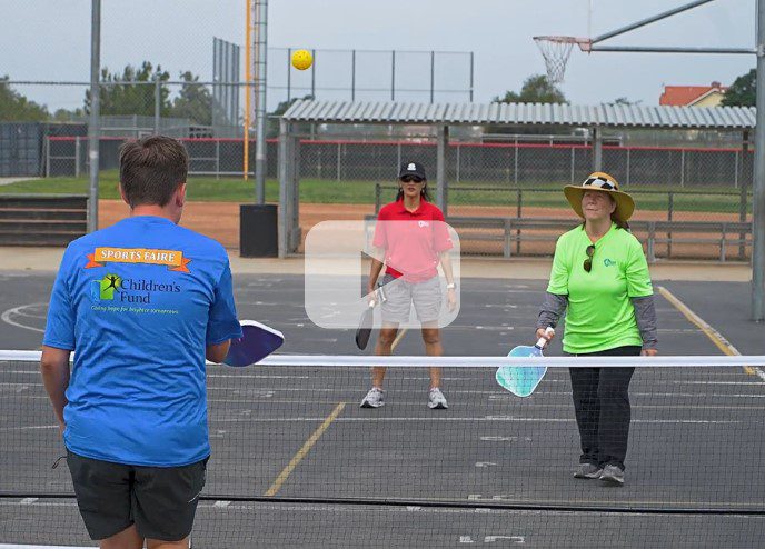 A youth and two women are playing pickleball outdoors. Image also includes a video play button overlay.
