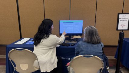 Two women sit at a table viewing a computer screen. One points at the screen while explaining. A sign nearby reads "Voter Registration Assistance.
