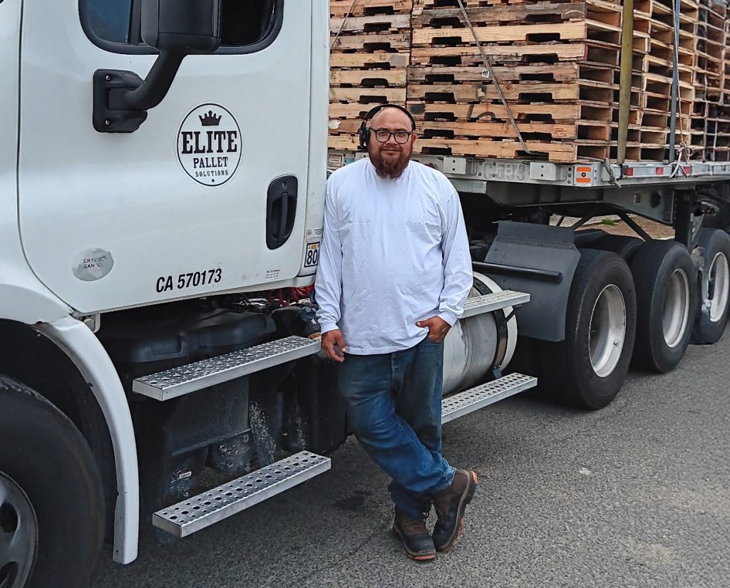 Man in white shirt and jeans stands next to a truck loaded with wooden pallets.