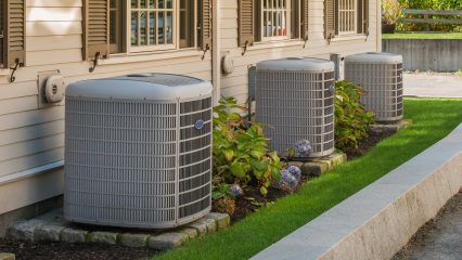 The side of a house features three air conditioning units and surrounding plants.