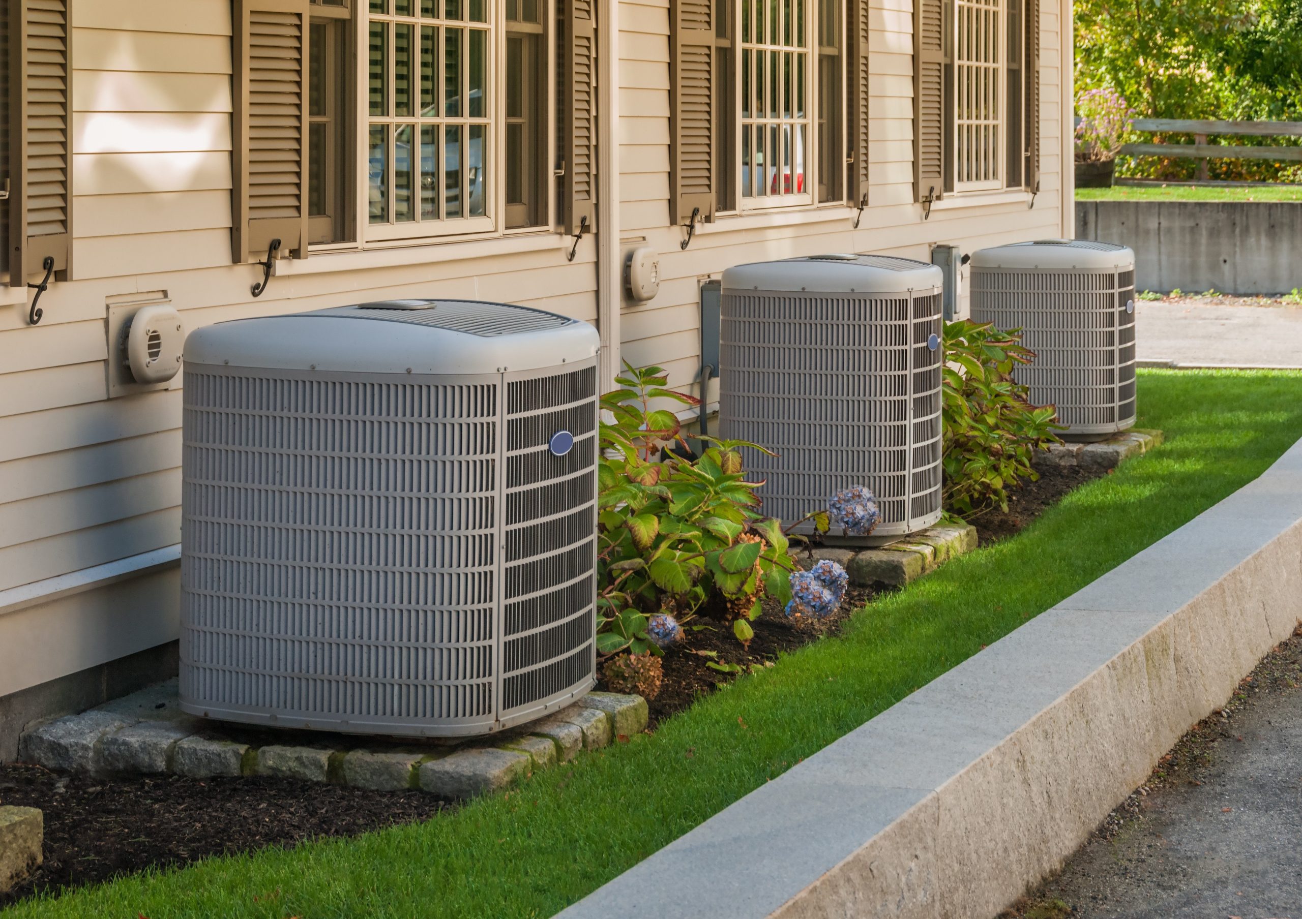 The side of a house features three air conditioning units and surrounding plants.