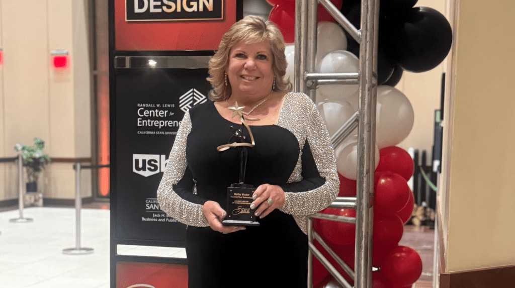 A smiling woman in a black dress holds a star-shaped award while standing beside red, white and black balloons at an event.