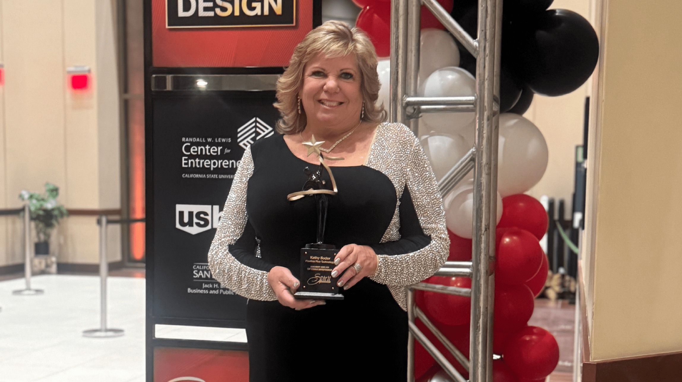 A smiling woman in a black dress holds a star-shaped award while standing beside red, white and black balloons at an event.