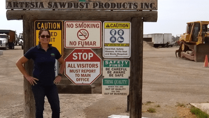 Person standing next to a sign for 'Artesia Sawdust Products Inc.', with cautionary notices and a piece of construction equipment in the background.