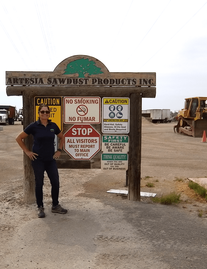 Person standing next to a sign for 'Artesia Sawdust Products Inc.', with cautionary notices and a piece of construction equipment in the background.