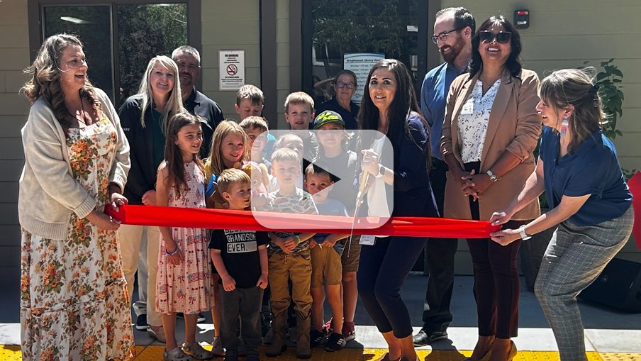 A group of adults and children smile at two women holding a red ribbon and another with scissors, with a play button overlay, indicating a video.