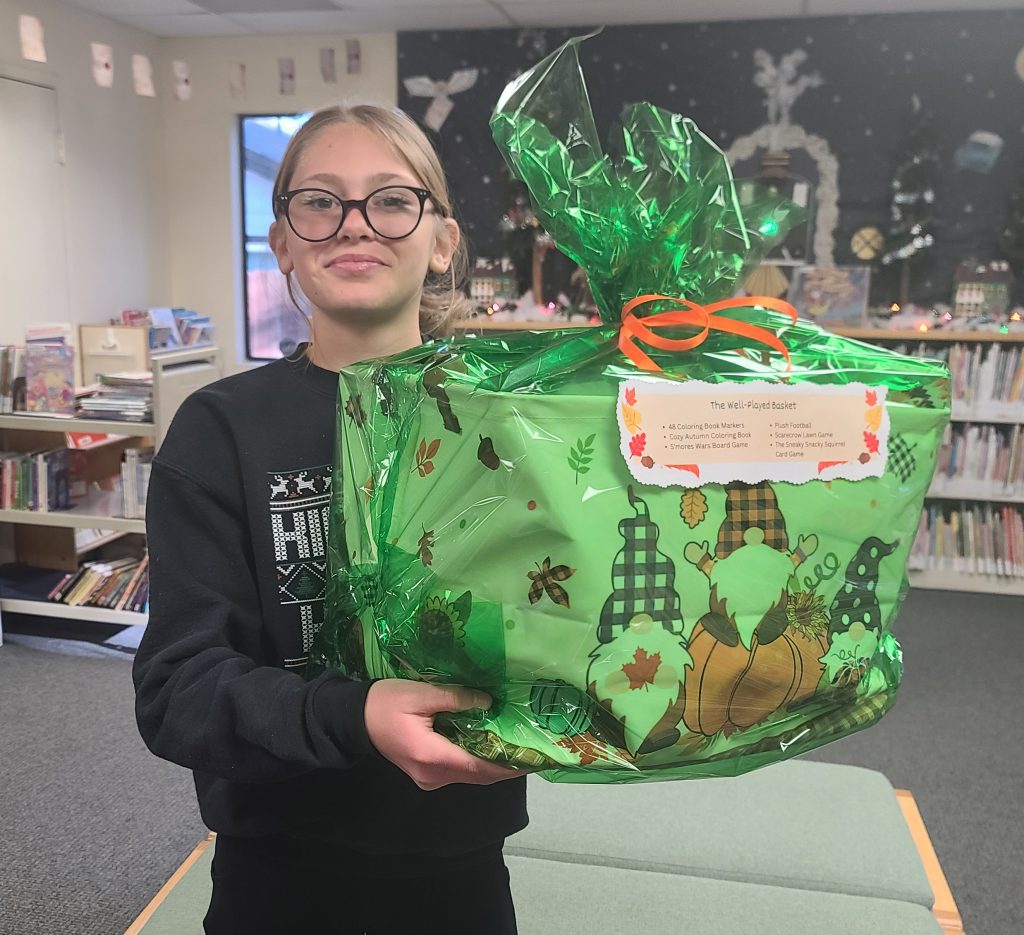 A person in glasses smiles while holding a large gift wrapped in green cellophane with orange ribbon.