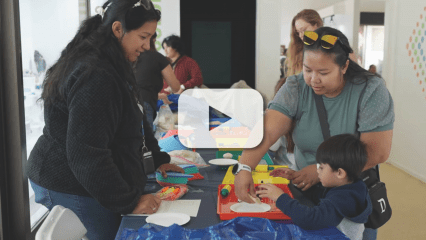 Two women assist a child with crafts at a table with colorful materials; other individuals in the background with video play button overlay.