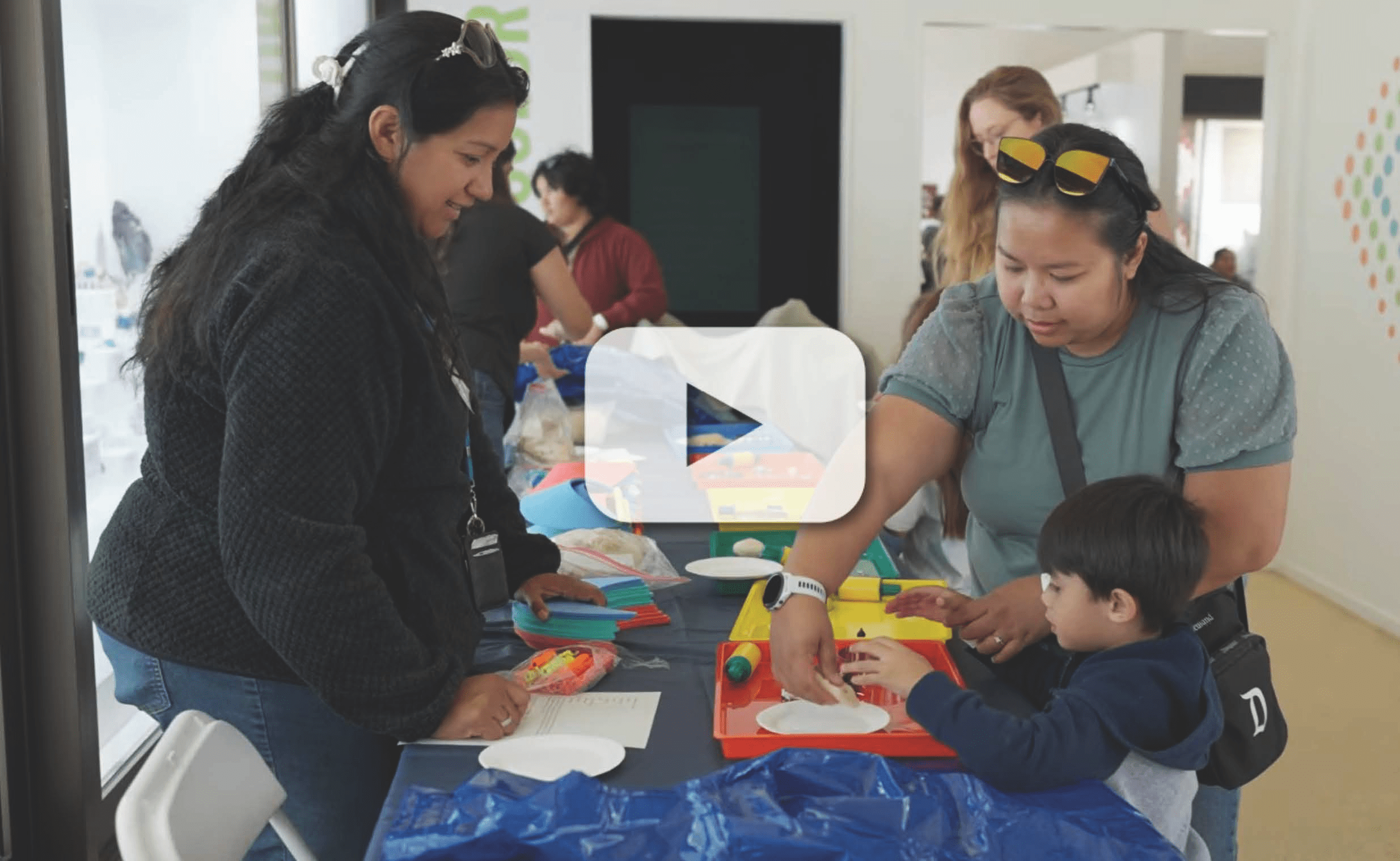 Two women assist a child with crafts at a table with colorful materials; other individuals in the background with video play button overlay.