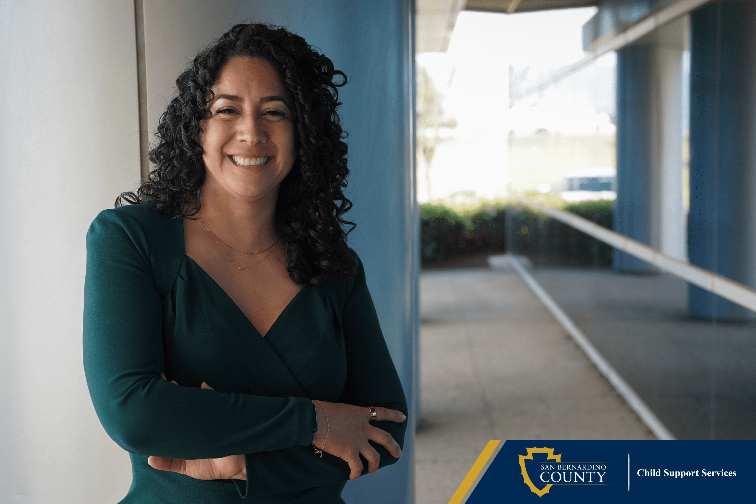 Woman standing outside near a pillar and window, with shrubs in the background and the Child Support Services logo in a blue and gold box.