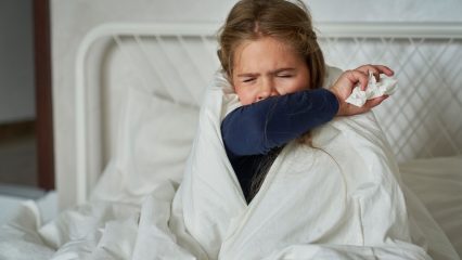 Child sitting up in bed, wrapped in a Child sitting up in bed, wrapped in a blanket, coughing into their arm while holding a tissue, illustrating respiratory illness symptoms.