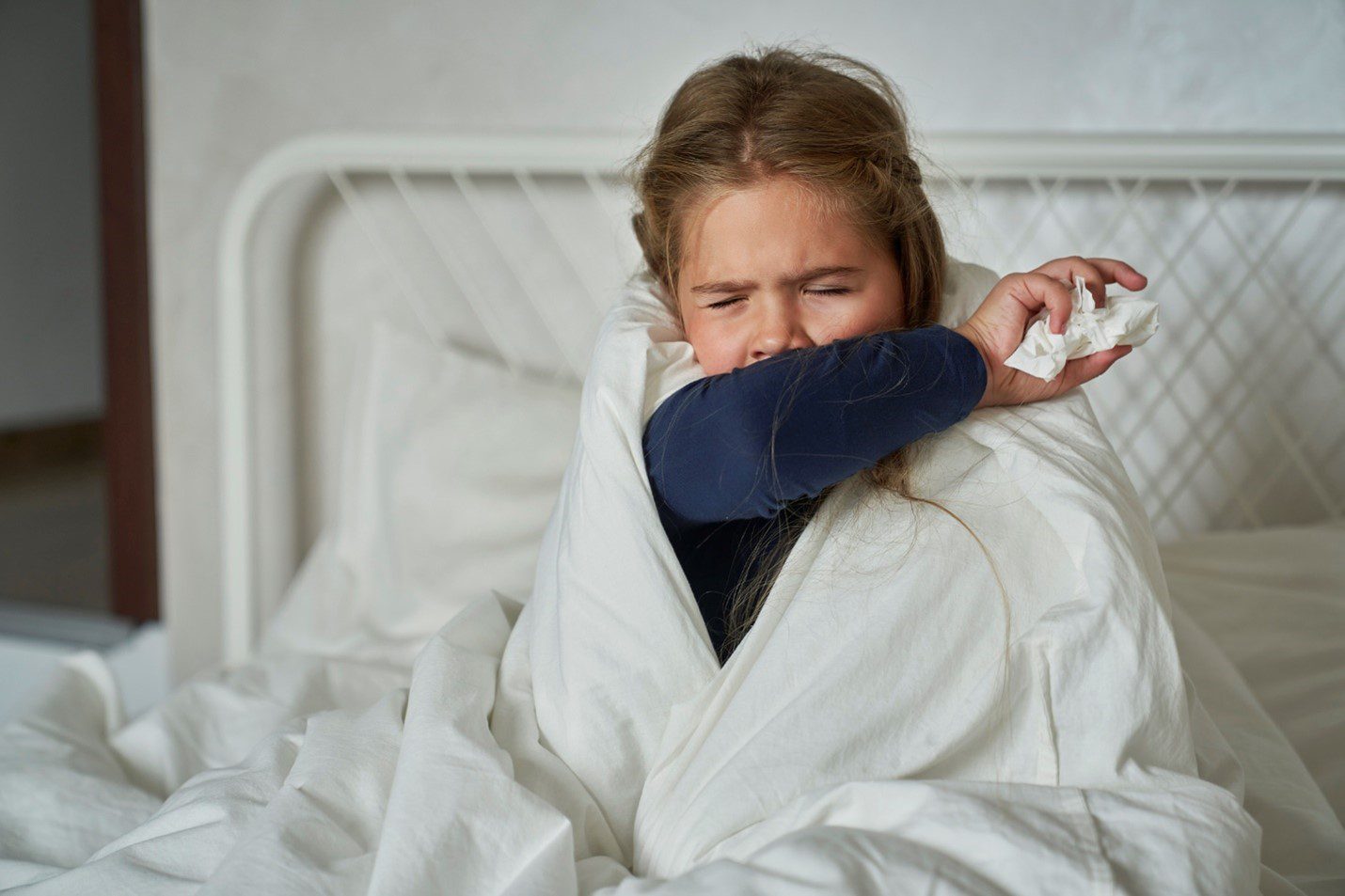 Child sitting up in bed, wrapped in a blanket, coughing into their arm while holding a tissue, illustrating respiratory illness symptoms.