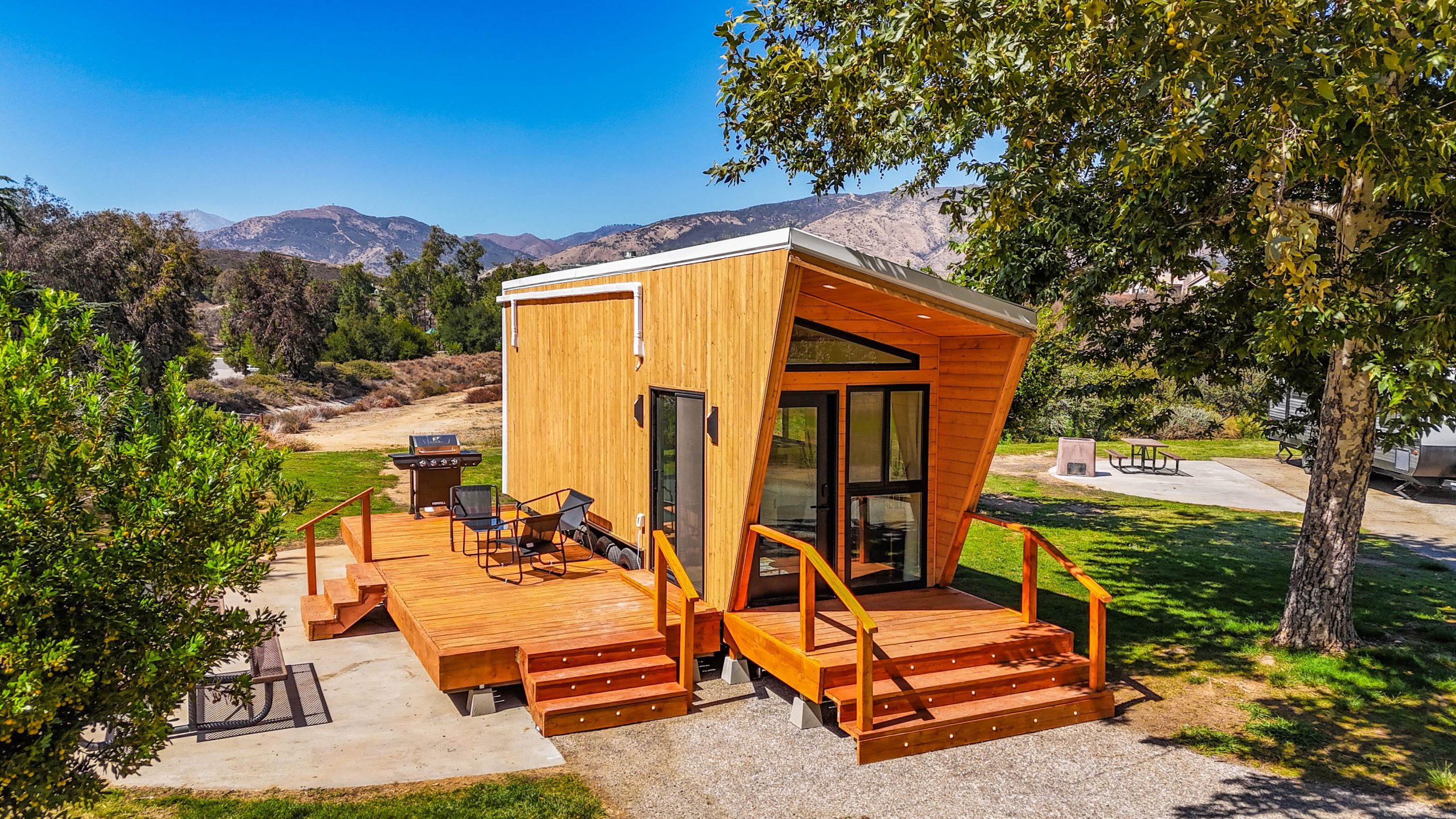 A wood cabin-style tiny home with a deck, barbecue, outdoor chairs, and stairs in a picturesque park setting with trees, grass and shrubs with a mountain background and blue sky.