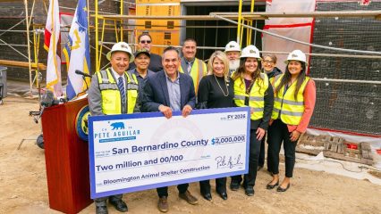 A group of smiling people, some in safety vests and helmets, stand holding a large check for $2 million. They are at a construction site with scaffolding.
