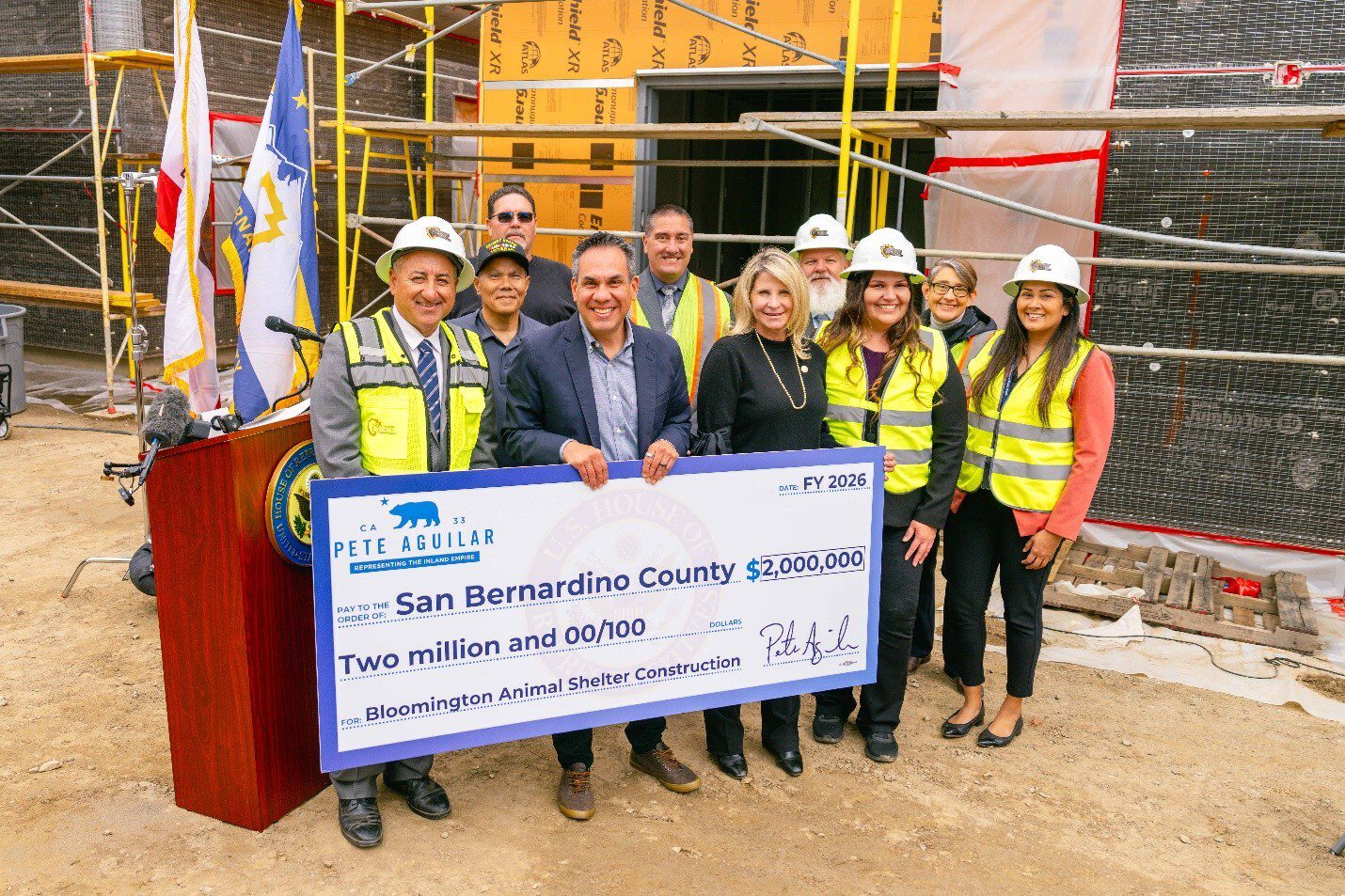 A group of smiling people, some in safety vests and helmets, stand holding a large check for $2 million. They are at a construction site with scaffolding.