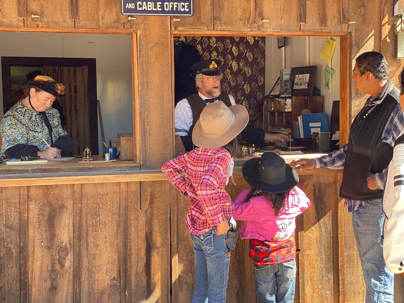 A man and woman dressed in 1800s period clothing stand at an old western telegraph office with a man and two children facing the man at the window of the office.