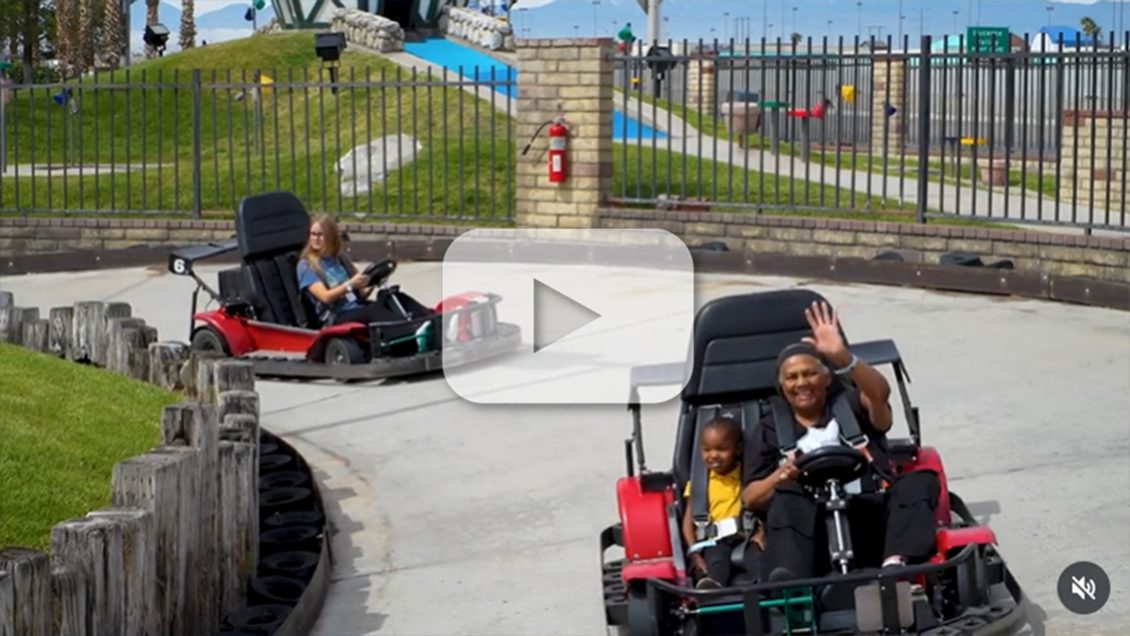 A woman is driving a go-cart at an amusement park and waiving hello with a child seated next to her and a teenager in a go-cart behind her.