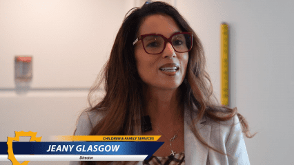 Jeany Glasgow, Director of Children & Family Services, speaks indoors, wearing red glasses and a light blazer with a microphone clipped on.