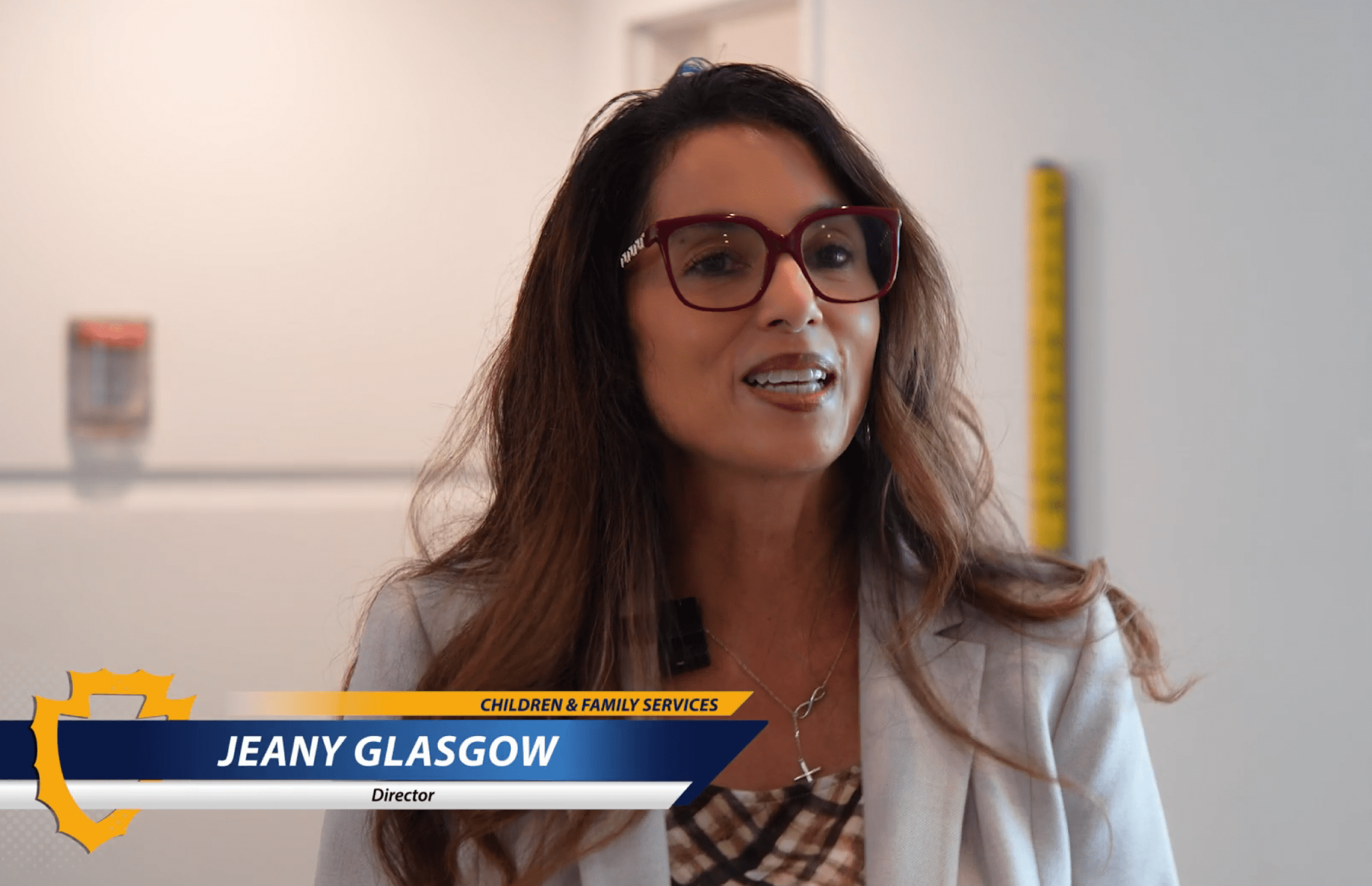 Jeany Glasgow, Director of Children & Family Services, speaks indoors, wearing red glasses and a light blazer with a microphone clipped on.