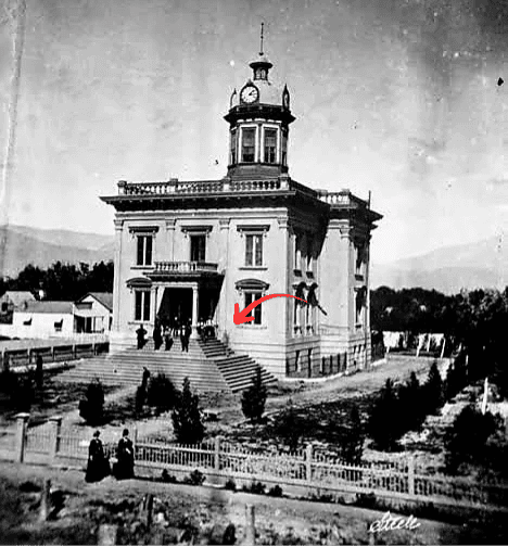 Black and white photo of the county courthouse featuring a clock tower and surrounded by a fence and small trees. Figures stand on the steps leading to the entrance.
