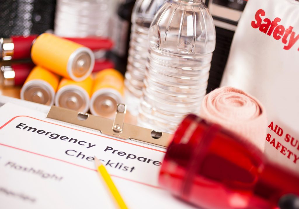 Emergency preparedness items including water bottles, batteries, a first aid kit and a red flashlight. A checklist on a clipboard is also shown.