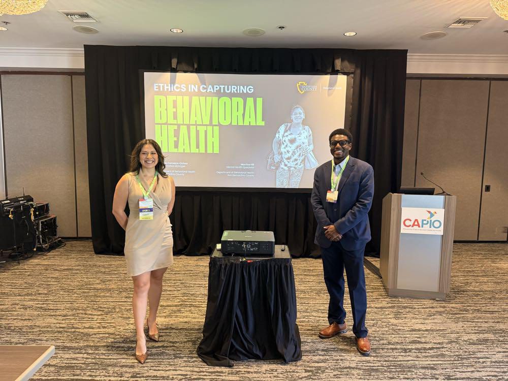 Two presenters stand next to each other in a conference room in front of a presentation with a slide reading “Ethics in Capturing Behavioral Health.”
