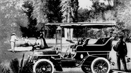 A vintage car is parked outdoors and surrounded by trees and several individuals.