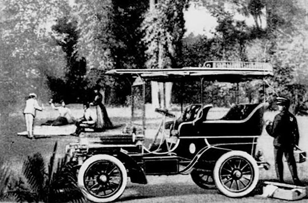 A vintage car is parked outdoors and surrounded by trees and several individuals.