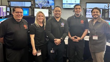 Five individuals from ICEMA and CONFIRE pose inside a busy emergency dispatch center with several computer and tv monitors.