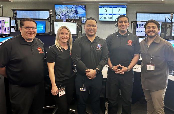 Five individuals from ICEMA and CONFIRE pose inside a busy emergency dispatch center with several computer and tv monitors.