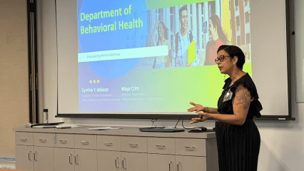 A woman in a black dress presents in a well-lit room, standing beside a large screen displaying a "Department of Behavioral Health" slide.