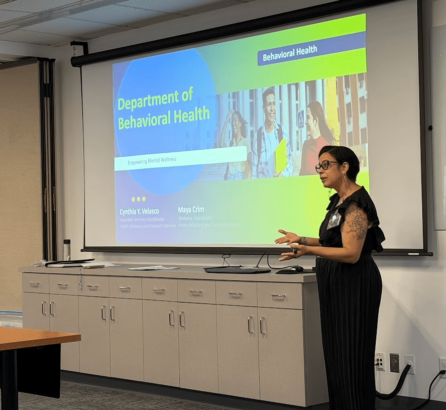A woman in a black dress presents in a well-lit room, standing beside a large screen displaying a "Department of Behavioral Health" slide.