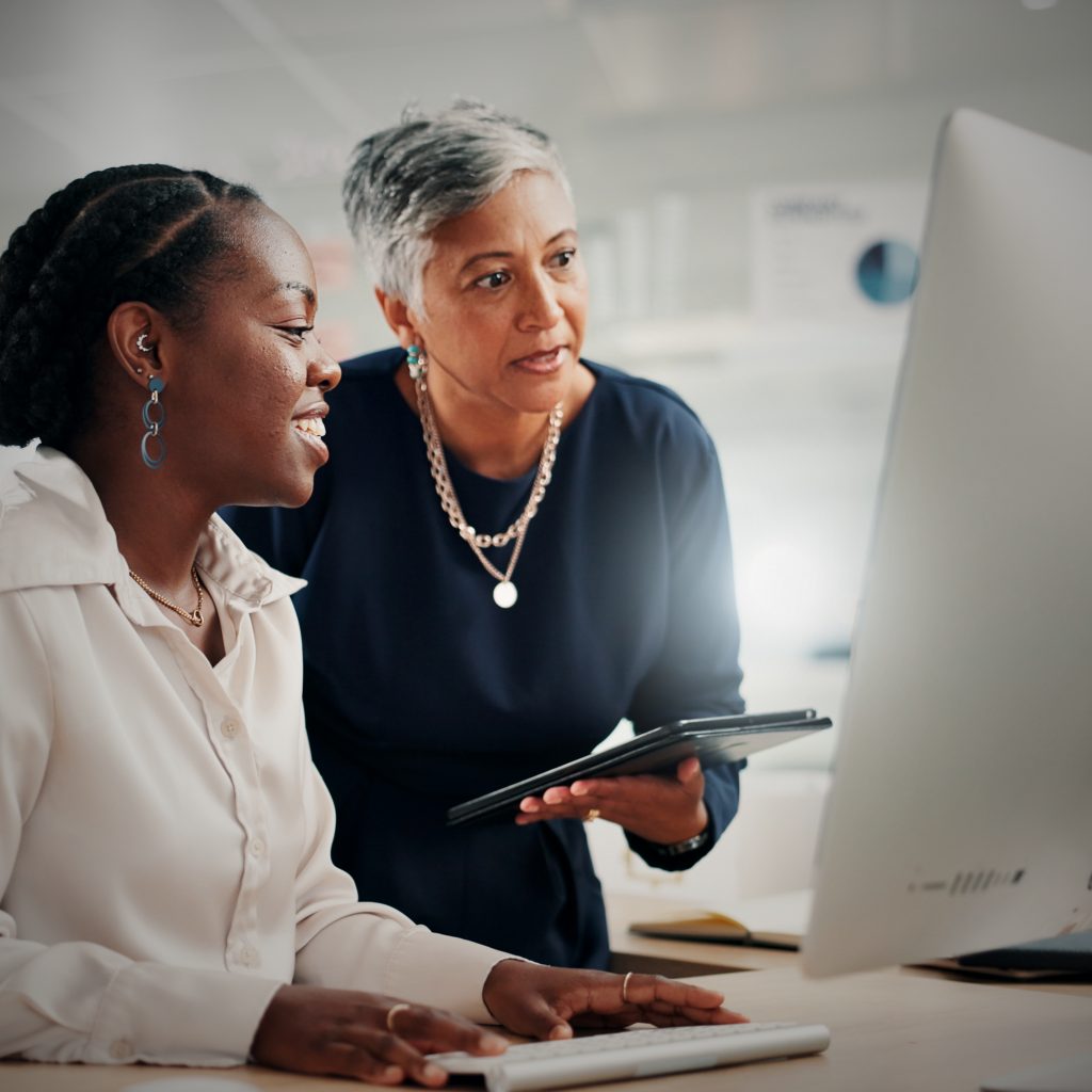 Two coworkers collaborate at a desk, with one person pointing at a computer monitor while the other types, discussing work in a modern office setting.