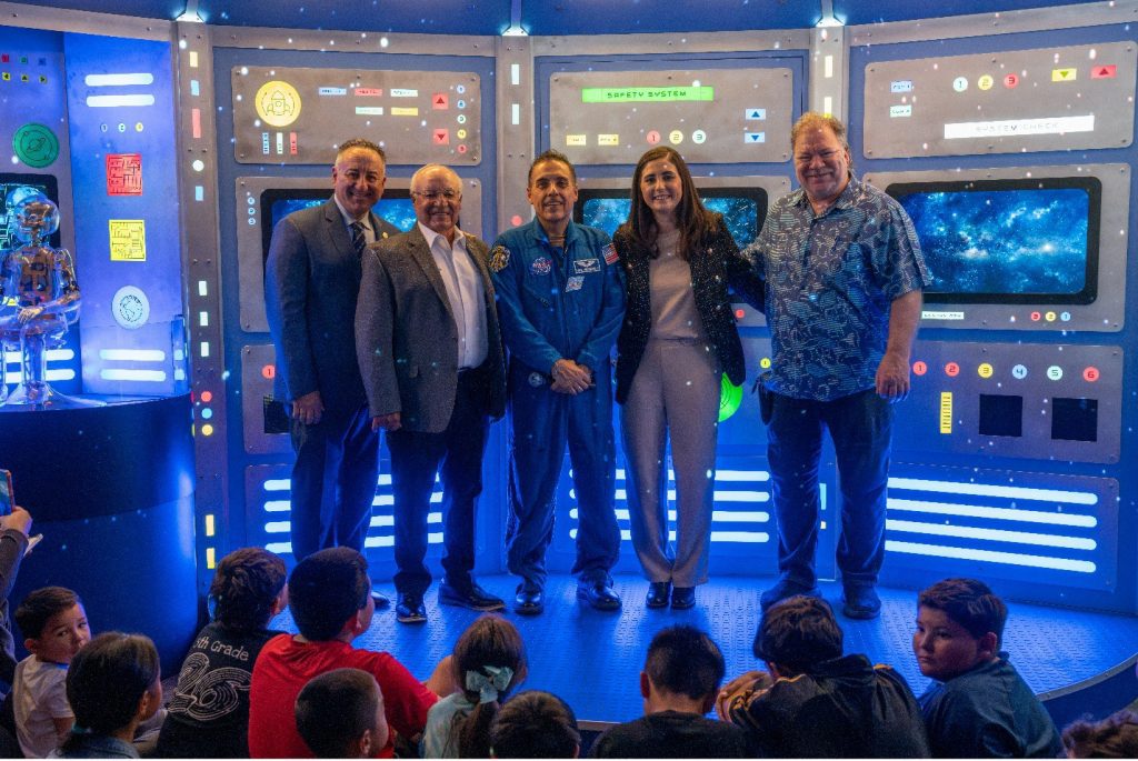 Five adults, including a uniformed astronaut, smile in a glowing sci-fi control room while children watch nearby at an upbeat space event.
