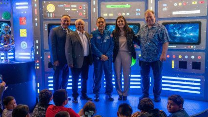 Five adults, including a uniformed astronaut, smile in a glowing sci-fi control room while children watch nearby at an upbeat space event.