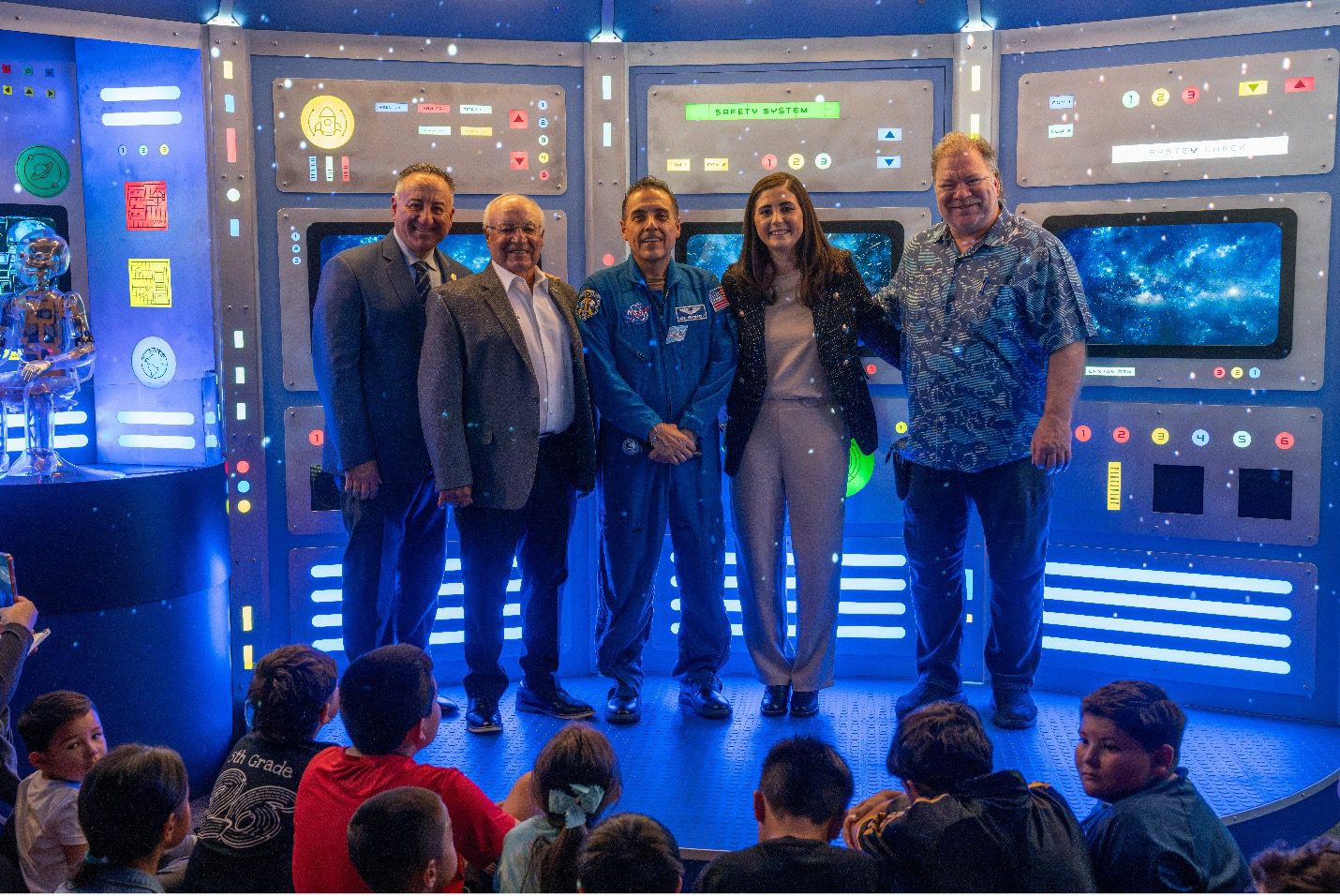 Five adults, including a uniformed astronaut, smile in a glowing sci-fi control room while children watch nearby at an upbeat space event.