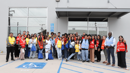 A diverse group of smiling people stands outside a building wearing colorful vests. Some hold blue tote bags.