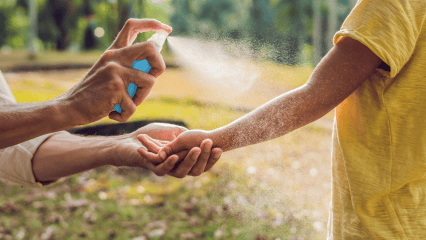 An adult sprays insect repellent onto a child’s arm outdoors in a sunny, grassy area.
