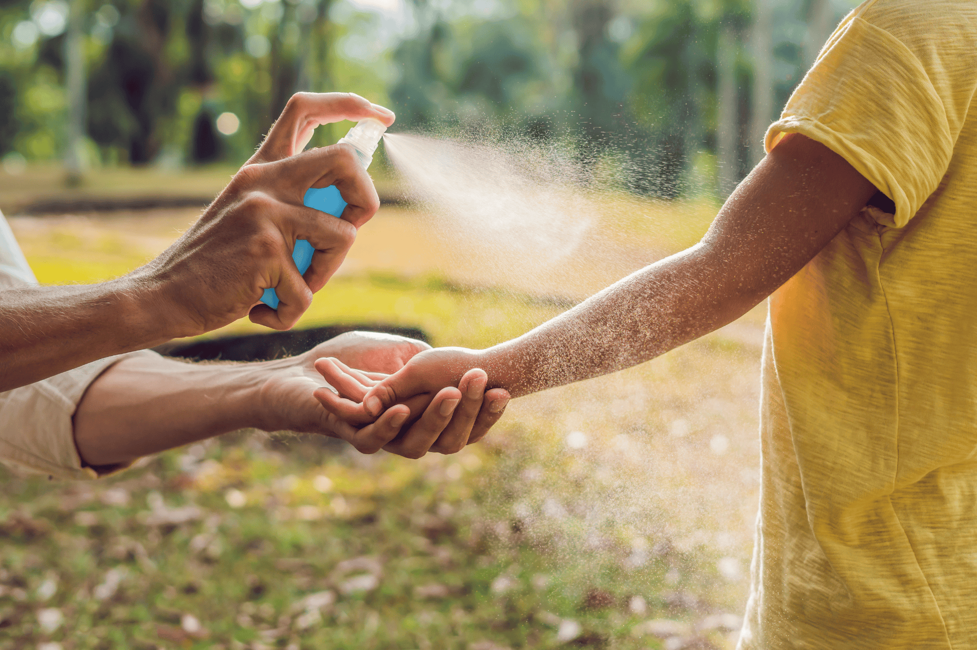 An adult sprays insect repellent onto a child’s arm outdoors in a sunny, grassy area.