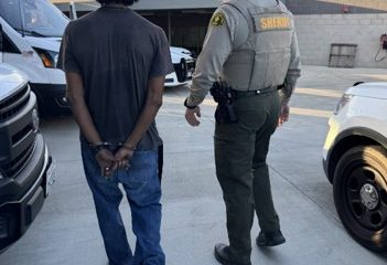 Sheriff escorts a handcuffed person through a parking area between patrol vehicles.