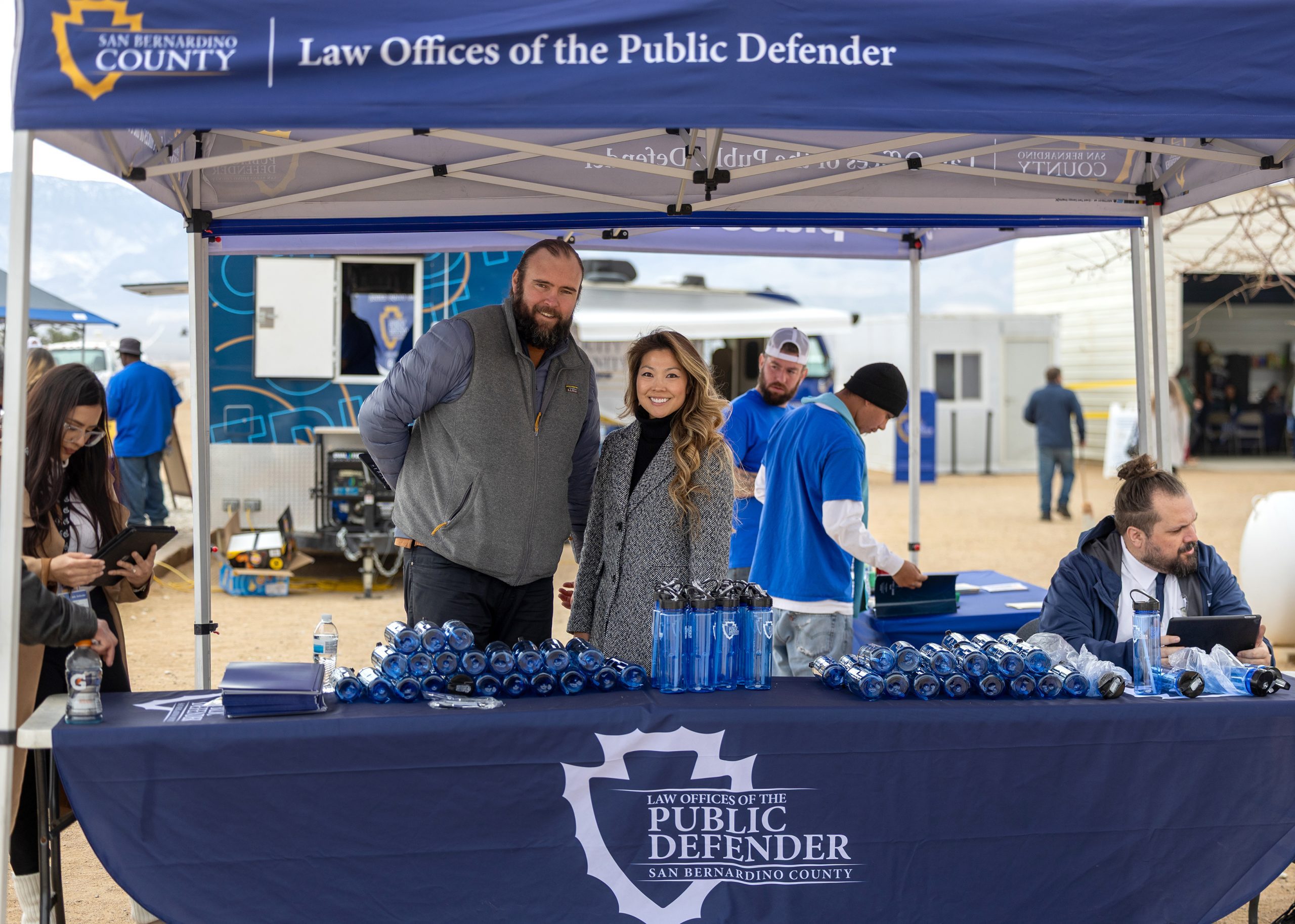 Several individuals stand behind a booth for the Law Offices of the San Bernardino County Public Defender as they give out blue water bottles.