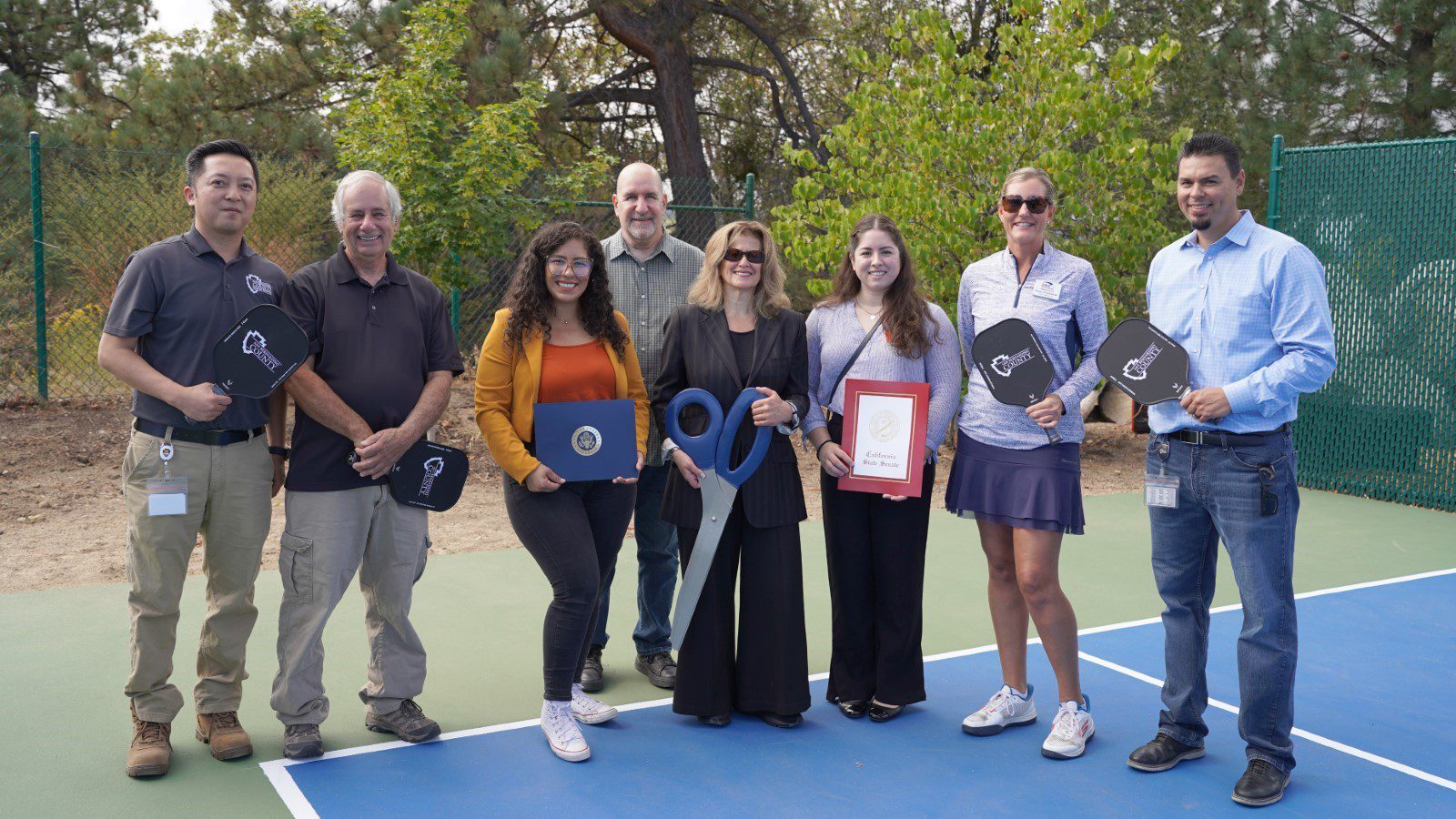 Eight people pose on a pickleball court holding paddles, certificates, and oversized scissors for a ribbon-cutting event.