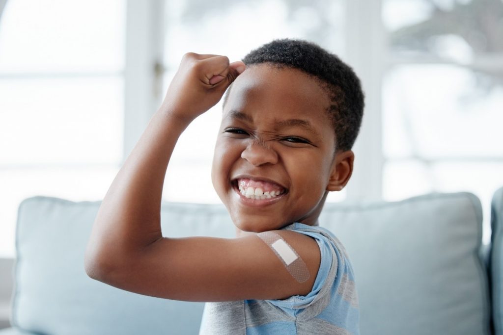 Smiling young child flexing their arm with a small bandage on the upper arm after receiving a vaccination.