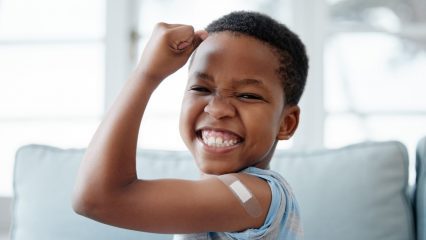 Smiling young child flexing their arm with a small bandage on the upper arm after receiving a vaccination.