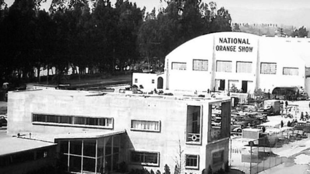 Black and white image of the National Orange Show event center with parked cars and surrounding landscape.