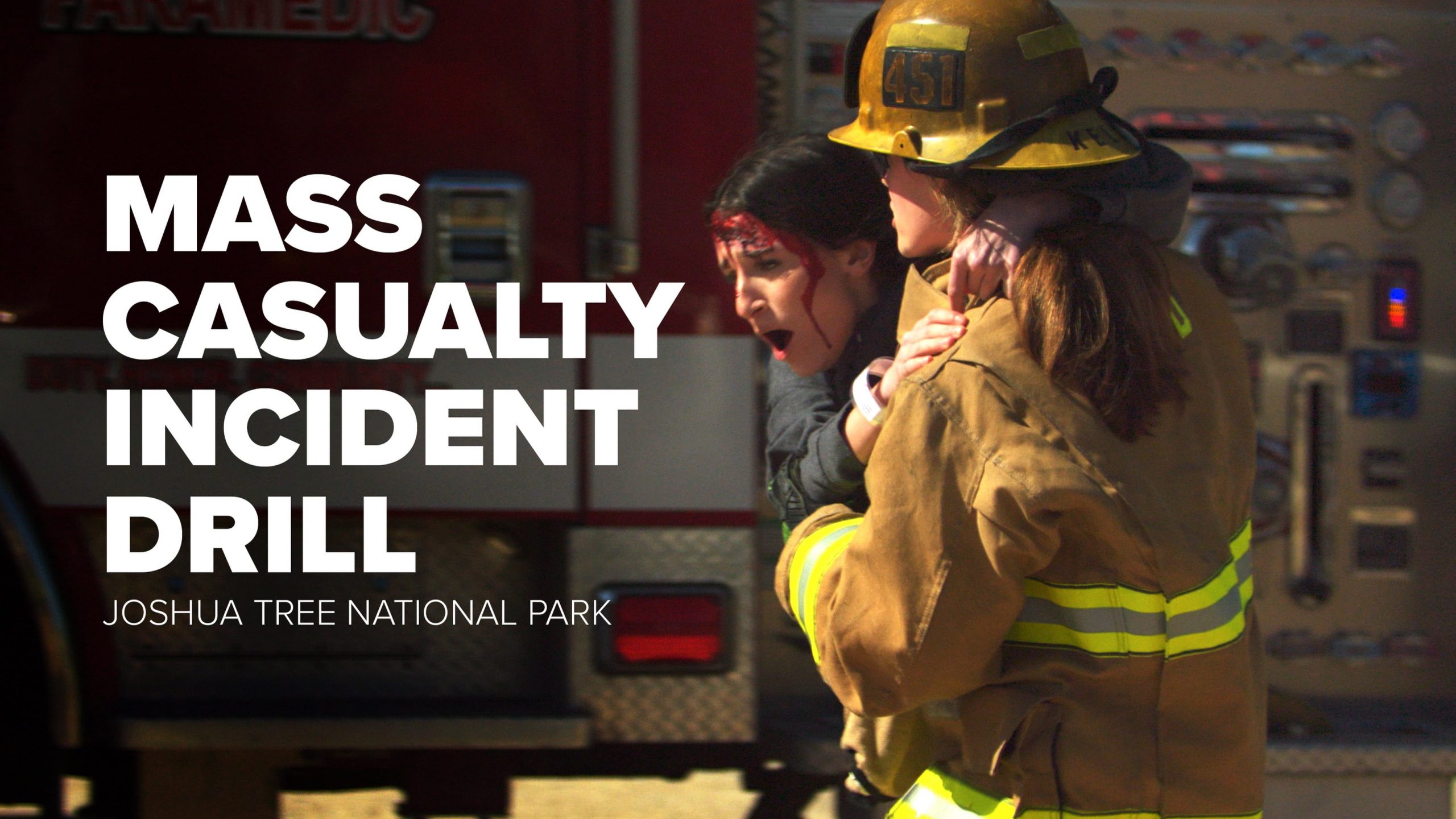 A firefighter in full turnout gear carries a distressed woman with simulated head injuries during a mass casualty incident drill at Joshua Tree National Park. A fire engine is visible in the background with text that says, “Mass Casualty Incident Drill Joshua Tree National Park.”