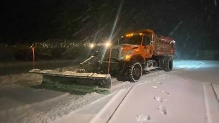 County snowplow clears a snow-covered mountain road at night during an active winter storm.
