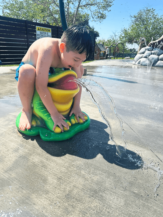 A young boy in swim trunks mimics a frog water feature with water coming out of its mouth at the splash pad at Prado Regional Park.